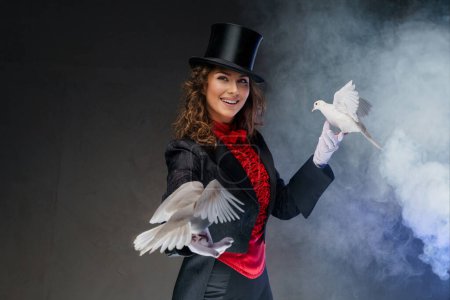 Ein bezaubernder Moment als Zauberin im Zaubererkostüm und schwarzem Zylinderhut zeigt bezaubernde Zaubertricks mit anmutigen weißen Tauben vor dunklem, rauchigen Hintergrund.