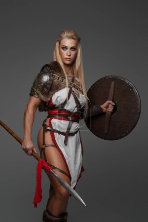 Foto de Mujer vikinga, vestida con armadura de fantasía con muslos desnudos. Ella con confianza empuña un escudo y lanza sobre un fondo gris - Imagen libre de derechos