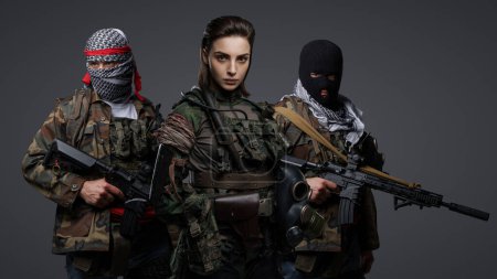 Téléchargez les photos : Groupe de trois militants du Moyen-Orient vêtus d'uniformes de camouflage, de keffiyehs et de cagoules posant sur un fond gris - en image libre de droit