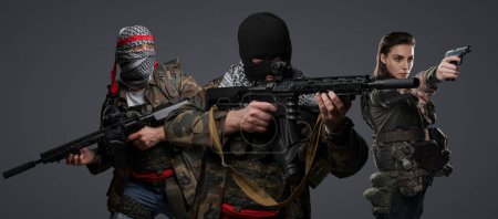 Foto de Trío de extremistas radicales de Oriente Medio disfrazados de camuflaje, keffiyeh y pasamontañas, posando sobre un fondo gris - Imagen libre de derechos