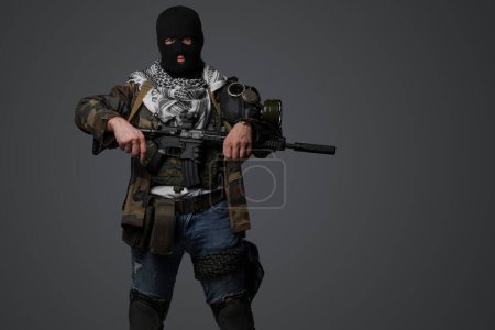 Téléchargez les photos : Portrait d'un militant du Moyen-Orient, vêtu d'une tenue noire de cagoule et de champ de camouflage, tenant un fusil, sur fond gris - en image libre de droit