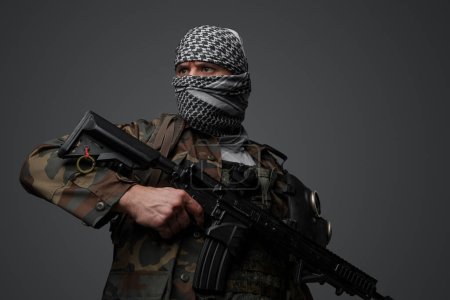Téléchargez les photos : Militant du Moyen-Orient, vêtu d'un keffiyeh blanc et d'une tenue de campagne camouflée, armé d'un fusil automatique, sur fond de gris neutre - en image libre de droit