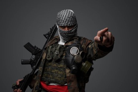 Foto de Soldado fanático radical de Oriente Medio, vestido con un keffiyeh blanco y camuflado uniforme de campo, armado con un rifle, apuntando con su dedo amenazadoramente contra un fondo gris - Imagen libre de derechos