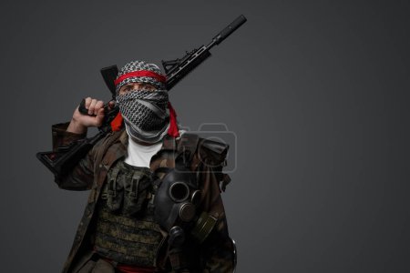 Photo for Middle Eastern radical fanatic soldier donning a white keffiyeh and camouflaged field uniform, armed with an automatic rifle, against a gray background - Royalty Free Image
