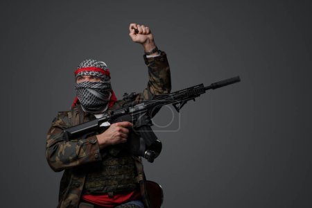 Photo for Fanatic soldier from the Middle East, wearing a white keffiyeh and camouflaged field uniform, armed with a rifle, posing with a raised hand against a gray background - Royalty Free Image