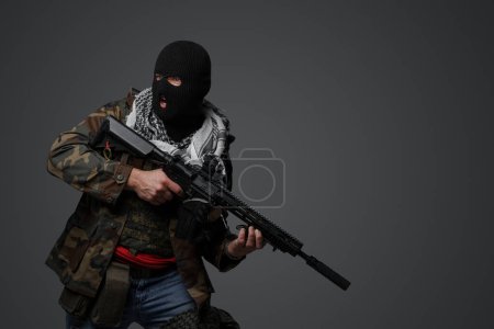 Foto de Un soldado radical de Oriente Medio vestido con pasamontañas negro y camuflado uniforme de campo, armado con un rifle, sobre un fondo gris - Imagen libre de derechos