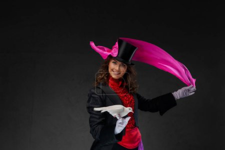 Téléchargez les photos : Une illustratrice élégante, vêtue d'un costume de magicien et d'un chapeau de cylindre noir, effectue des tours envoûtants avec une colombe blanche gracieuse et un mouchoir en soie violette sur fond sombre - en image libre de droit