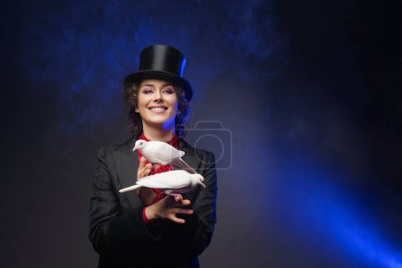 Téléchargez les photos : Une magicienne joyeuse, vêtue d'un costume magique et d'un chapeau noir, effectue des tours enchanteurs avec des colombes blanches sur un fond sombre éclairé par la lumière bleue - en image libre de droit
