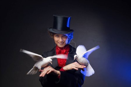Photo for A cheerful female magician, dressed in a magical costume and a black top hat, performs enchanting tricks with white doves against a dark background illuminated by blue light - Royalty Free Image