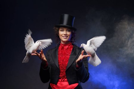Foto de Una alegre maga, vestida con un traje mágico y un sombrero de copa negro, realiza encantadores trucos con palomas blancas sobre un fondo oscuro iluminado por la luz azul - Imagen libre de derechos