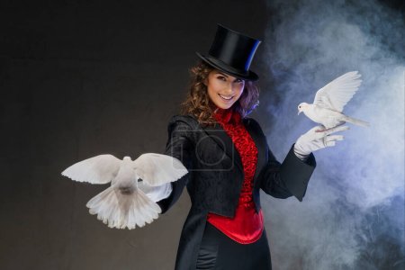 Ein bezaubernder Moment als Zauberin im Zaubererkostüm und schwarzem Zylinderhut zeigt bezaubernde Zaubertricks mit anmutigen weißen Tauben vor dunklem, rauchigen Hintergrund.