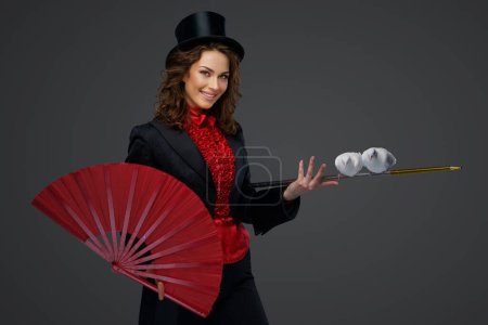 Téléchargez les photos : Joyeux illusionniste féminin effectuant des tours de magie avec des colombes blanches tout en tenant un grand ventilateur rouge sur un fond gris. - en image libre de droit
