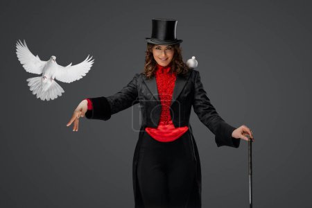 Téléchargez les photos : Elégant illusionniste en costume et chapeau de cylindre noir effectue magie de colombe enchanteresse sur un fond gris tamisé - en image libre de droit