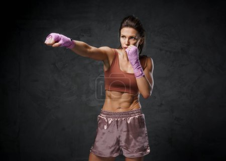 Foto de Mujer musculosa boxeadora demostrando habilidades sorprendentes, vestida con atuendo deportivo, contra un fondo oscuro malhumorado - Imagen libre de derechos