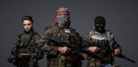 Foto de Grupo de tres militantes de Oriente Medio vestidos con uniformes de camuflaje, keffiyehs y pasamontañas posando sobre un fondo gris - Imagen libre de derechos