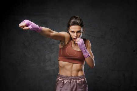 Foto de Atlético luchador femenino en sujetador deportivo y pantalones cortos que muestran la técnica de punch con puños envueltos en un fondo oscuro - Imagen libre de derechos
