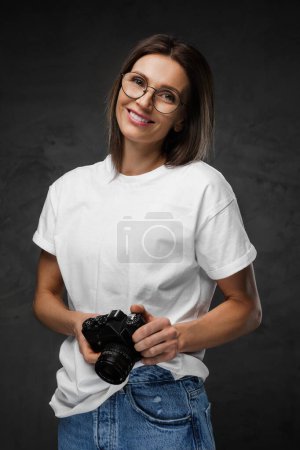 Foto de Mujer alegre en gafas y una camiseta blanca sosteniendo una cámara sobre un fondo oscuro - Imagen libre de derechos