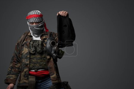 Photo for Militant from the Middle East, donned in a white keffiyeh and camouflaged field attire, wielding a rifle, posing with a black balaclava held in hand against a gray backdrop - Royalty Free Image