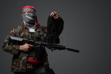 Téléchargez les photos : Militant du Moyen-Orient, revêtu d'un keffiyeh blanc et d'une tenue de campagne camouflée, brandissant un fusil, posant avec une cagoule noire tenue à la main sur un fond gris - en image libre de droit