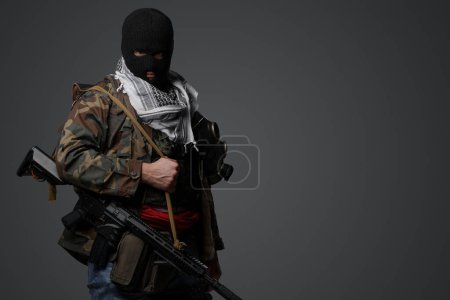 Téléchargez les photos : Soldat radical du Moyen-Orient vêtu d'une cagoule noire et d'un uniforme de campagne camouflé, armé d'un fusil, sur fond gris - en image libre de droit