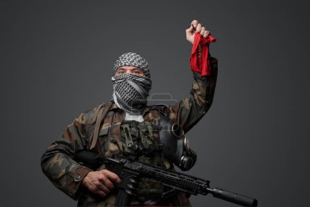 Photo for Middle Eastern radical fanatic soldier, dressed in a white keffiyeh and camouflaged field uniform, armed with a rifle, raised hand holding a red cloth and a remote control - Royalty Free Image