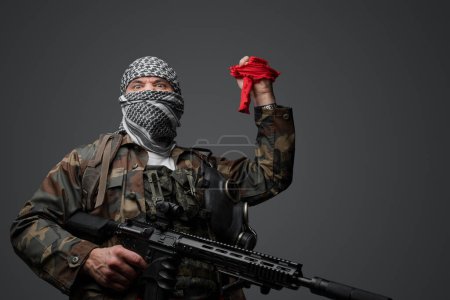 Photo for Middle Eastern radical fanatic soldier, dressed in a white keffiyeh and camouflaged field uniform, armed with a rifle, raised hand holding a red cloth and a remote control - Royalty Free Image