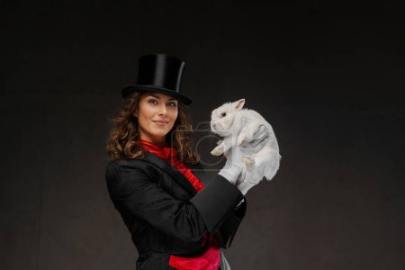 Téléchargez les photos : Une magicienne envoûtante, vêtue d'un costume de magicien et d'un chapeau noir, effectue des tours enchanteurs avec un charmant lapin blanc sur fond sombre - en image libre de droit