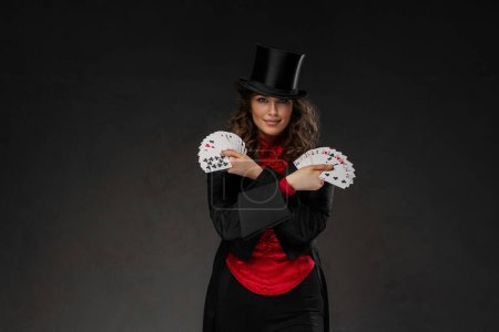 Foto de Encantadora mago femenino en un disfraz de magos y sombrero de copa negro realizar trucos de cartas sobre un fondo oscuro - Imagen libre de derechos
