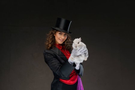 Foto de Una mago fascinante, vestida con un disfraz de magos y un sombrero de copa negro, realiza trucos encantadores con un encantador conejo blanco contra un fondo oscuro - Imagen libre de derechos