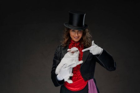 Foto de Una mago fascinante, vestida con un disfraz de magos y un sombrero de copa negro, realiza trucos encantadores con un encantador conejo blanco contra un fondo oscuro - Imagen libre de derechos