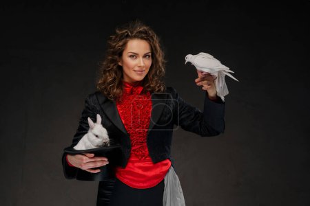 Téléchargez les photos : Portrait d'illusionniste en costume de magicien et chapeau noir exécutant des tours de magie avec des colombes blanches sur un fond sombre - en image libre de droit