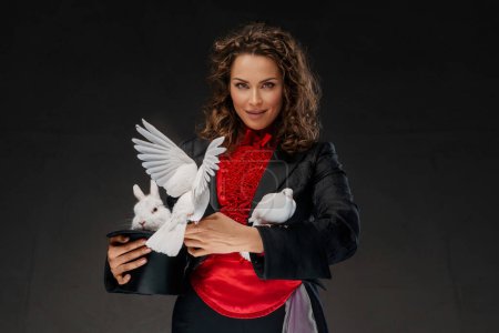 Téléchargez les photos : Portrait d'un magicien en costume de magicien et chapeau noir exécutant des tours de magie avec des colombes blanches sur un fond sombre - en image libre de droit