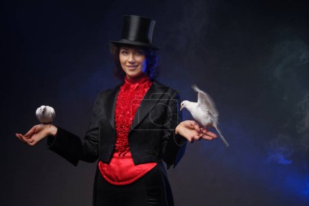Téléchargez les photos : Une magicienne joyeuse, vêtue d'un costume magique et d'un chapeau noir, effectue des tours enchanteurs avec des colombes blanches sur un fond sombre éclairé par la lumière bleue - en image libre de droit