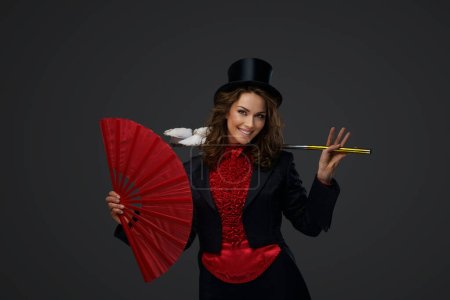Téléchargez les photos : Joyeux illusionniste féminin effectuant des tours de magie avec des colombes blanches tout en tenant un grand ventilateur rouge sur un fond gris. - en image libre de droit