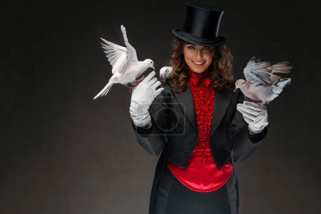 Téléchargez les photos : Portrait d'illusionniste en costume de magicien et chapeau noir exécutant des tours de magie avec des colombes blanches sur un fond sombre - en image libre de droit