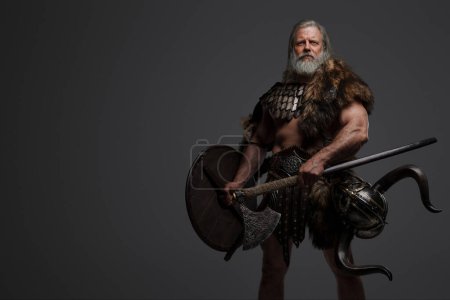 Foto de Anciano guerrero vikingo canoso, mostrando fuerza y sabiduría, vestido con pieles y armadura ligera, con un casco colgado en la cintura, blandiendo hachas dobles sobre un fondo neutro - Imagen libre de derechos