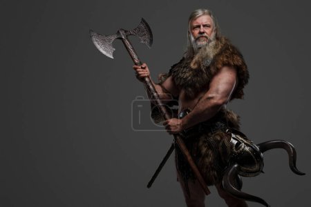 Foto de Anciano guerrero vikingo barbudo vestido con piel y armadura ligera, sosteniendo un hacha de dos manos, sobre un fondo gris - Imagen libre de derechos