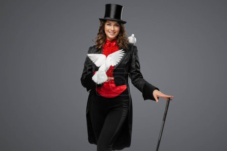 Téléchargez les photos : Elégant illusionniste en costume et chapeau de cylindre noir effectue magie de colombe enchanteresse sur un fond gris tamisé - en image libre de droit