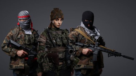 Foto de Grupo de tres militantes de Oriente Medio vestidos con uniformes de camuflaje, keffiyehs y pasamontañas posando sobre un fondo gris - Imagen libre de derechos