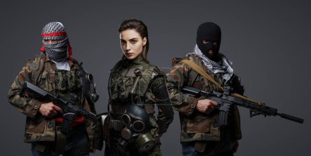 Foto de Trío de extremistas radicales de Oriente Medio disfrazados de camuflaje, keffiyeh y pasamontañas, posando sobre un fondo gris - Imagen libre de derechos