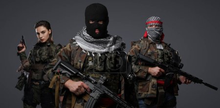 Photo for Group of three Middle Eastern militants dressed in camouflage uniforms, keffiyehs, and balaclavas posing against a gray background - Royalty Free Image