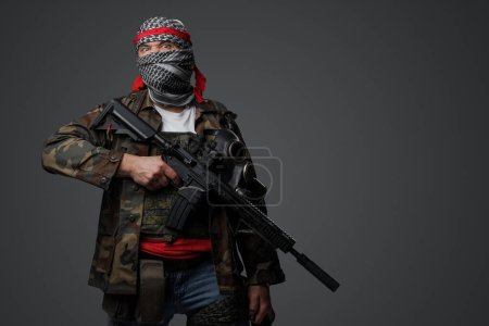 Téléchargez les photos : Soldat fanatique radical du Moyen-Orient revêtu d'un keffiyeh blanc et d'un uniforme de campagne camouflé, armé d'un fusil automatique, sur fond gris - en image libre de droit