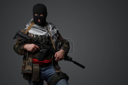 Photo for Portrait of a militant from the Middle East, wearing a black balaclava and camo field attire, holding a rifle, set against a gray backdrop - Royalty Free Image