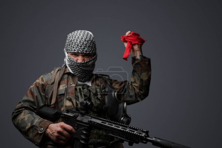Foto de Soldado fanático radical de Oriente Medio, vestido con un keffiyeh blanco y camuflado uniforme de campo, armado con un rifle, levantado de la mano sosteniendo una tela roja y un mando a distancia - Imagen libre de derechos
