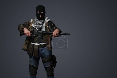 Foto de Retrato de un soldado radical de Oriente Medio vestido con un pasamontañas negro y un uniforme de campo camuflado, armado con un rifle automático, sobre un fondo gris frío - Imagen libre de derechos