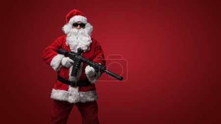 Téléchargez les photos : Un homme habillé comme le Père Noël, portant des lunettes de soleil noires élégantes, pose avec des mitrailleuses jouets sur un fond rouge audacieux - en image libre de droit