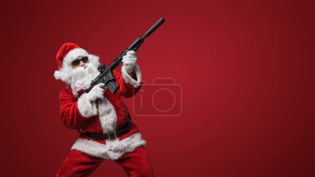 Foto de Un hombre vestido de Santa Claus, con elegantes gafas de sol negras, posa con ametralladoras de juguete sobre un atrevido telón de fondo rojo - Imagen libre de derechos