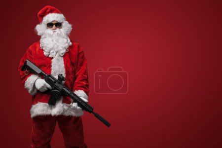 Foto de Un hombre vestido de Santa Claus, con elegantes gafas de sol negras, posa con ametralladoras de juguete sobre un atrevido telón de fondo rojo - Imagen libre de derechos