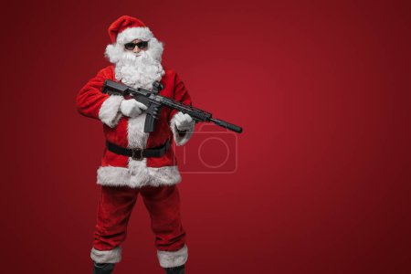 Photo for A man in a Santa Claus suit, wearing black sunglasses, poses with toy guns in hand against a red backdrop - Royalty Free Image