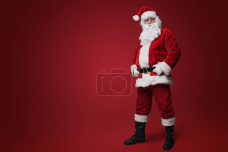 Téléchargez les photos : Joyeux Père Noël en costume rouge traditionnel debout avec les mains sur les hanches sur fond rouge - en image libre de droit